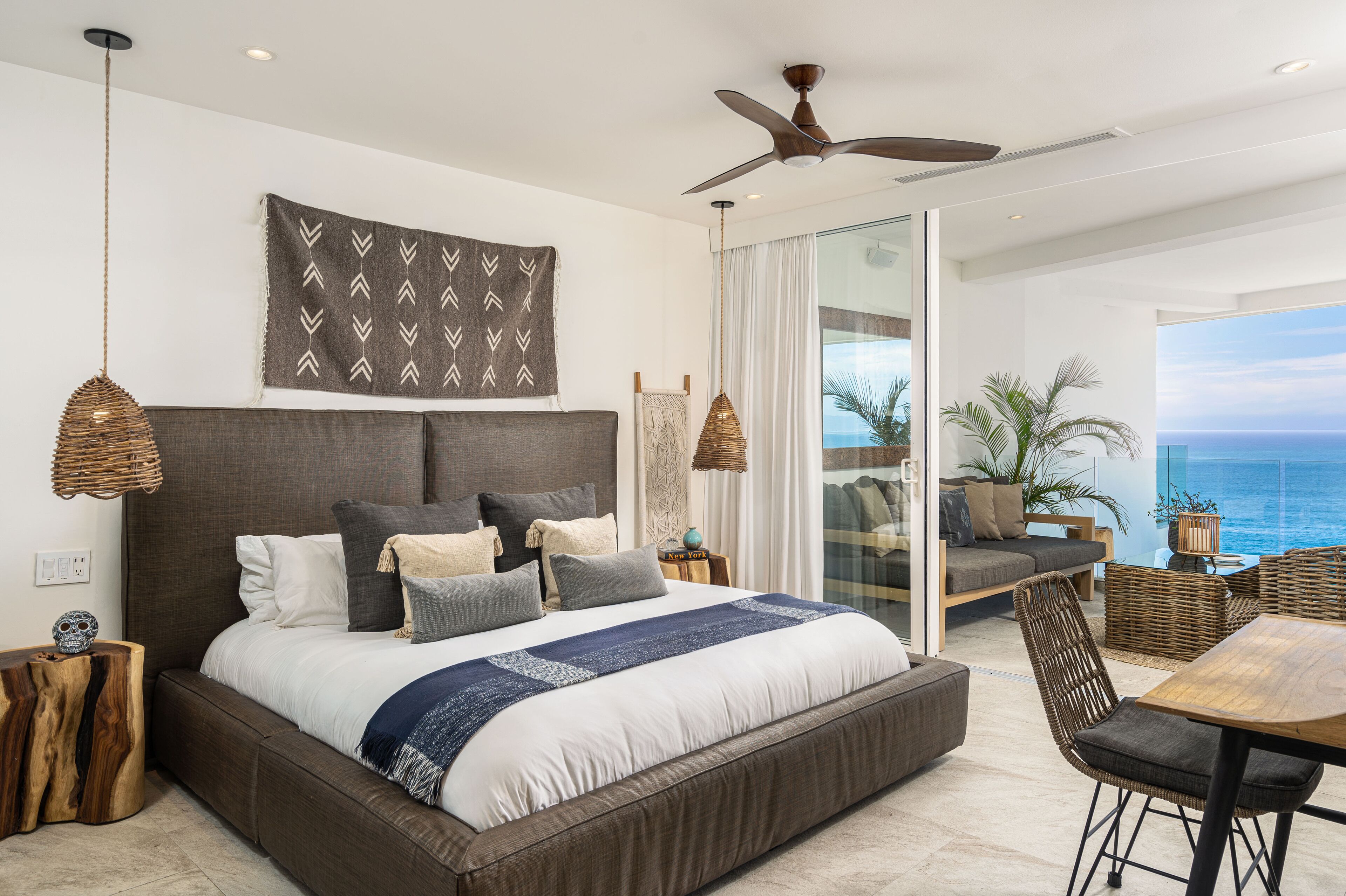 A modern bedroom with a large bed, a ceiling fan, and a view of the ocean.