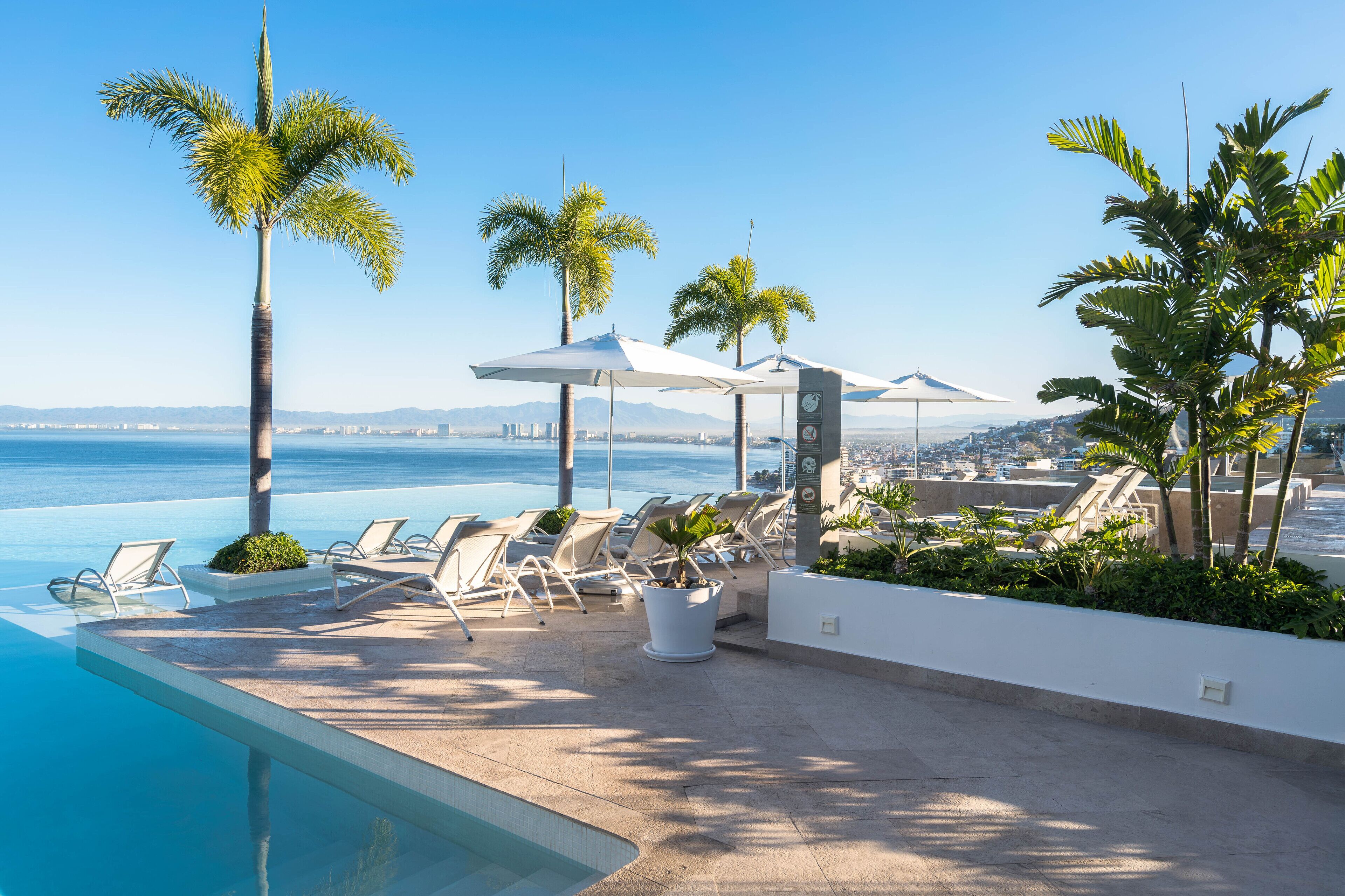 Zona junto a la piscina con sillones de descanso y sombrillas, con vista a una masa de agua y un horizonte urbano distante.