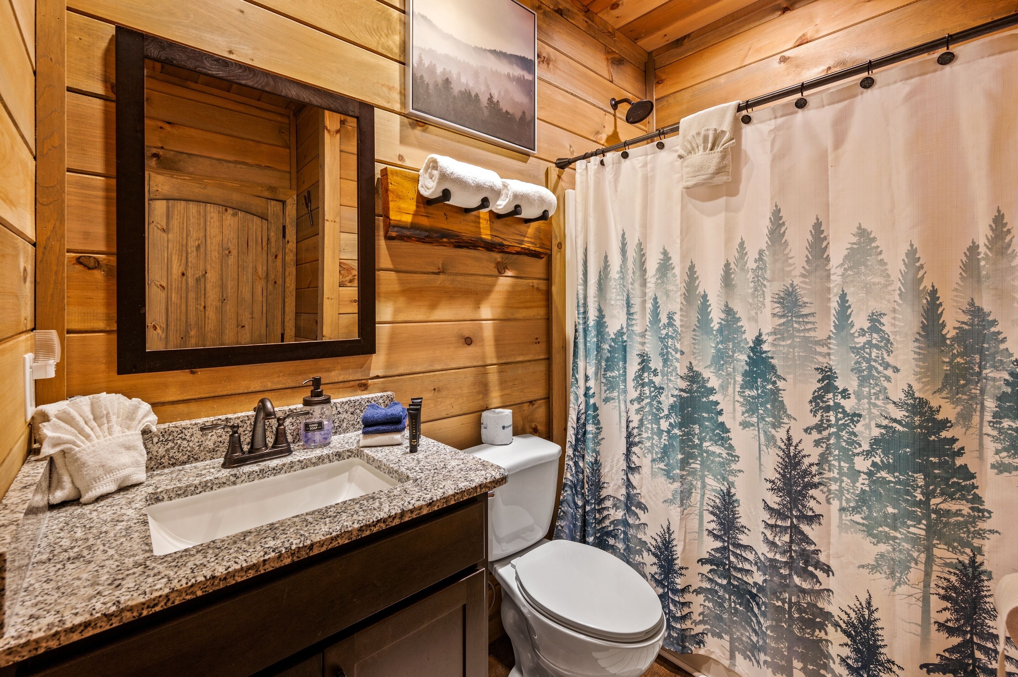 A wooden cabin bathroom with a granite sink, a toilet, and a shower curtain with a forest design.
