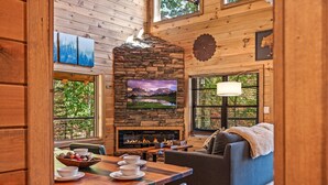 L’intérieur chaleureux d’un chalet avec une cheminée en pierre, un téléviseur fixé au-dessus, et un coin repas avec une table mise pour deux personnes.