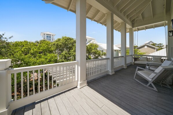 A covered outdoor deck with white railings, a wooden floor, and a seating area with a sofa and chairs.