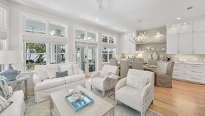 A spacious living room with a large window, a dining area, and a kitchen with white cabinetry. - Stella By The Sea 3 Bedroom Home by RedAwning (Panama City Beach)
