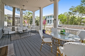 A covered outdoor patio with white wicker furniture, a glass table, and a view of a house with a white fence and palm trees.