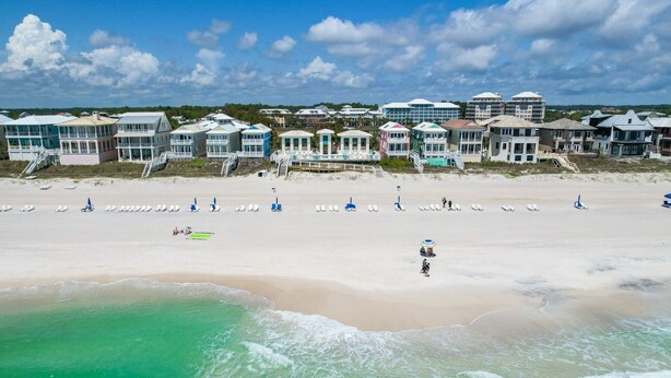 A beach with rows of beach chairs and umbrellas, a lifeguard tower, and a row of beachfront buildings. - Stella By The Sea 3 Bedroom Home by RedAwning (Panama City Beach)