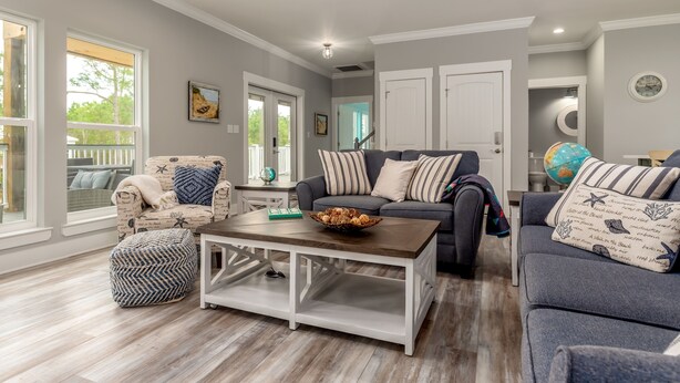A spacious living room with a large window, a dark grey sofa, a white coffee table, and a patterned ottoman. - Blue Haven - 1414 Oleander 4 Bedroom Home by RedAwning (Dauphin Island)