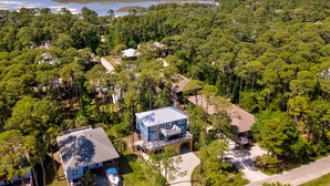An aerial view of a residential area with dense greenery, a large house with a blue roof, and a dock extending into a body of water. - Blue Haven - 1414 Oleander 4 Bedroom Home by RedAwning (Dauphin Island)