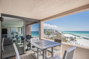 Uma varanda com mesa e cadeiras, com vista para uma praia com guarda-sóis e uma piscina.