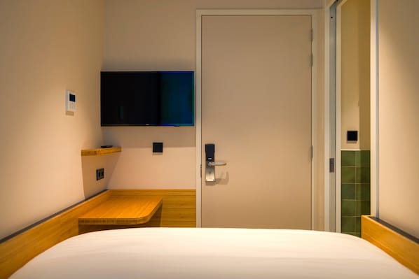 A hotel room with a bed, a mounted television, a wooden bench, a door, and a small shelf. - Poppies (Amsterdam)