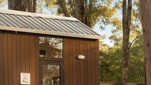 Una cabaña de madera con techo metálico, una puerta y un ventanal pequeño. Hay un banco de madera y una mesa con sillas al frente.