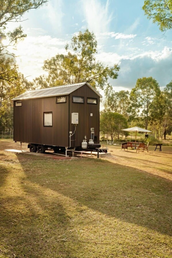 A small, single-story wooden cabin with a metal roof, a small porch, and a picnic table in a grassy area with trees in the background.
