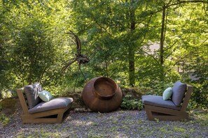 Twee houten stoelen met kussens, een grote decoratieve pot en een spinnenbeeld in een tuin.