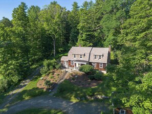 A two-story house with a stone foundation, surrounded by dense forest, featuring a front porch and a visible staircase leading to the entrance. - Classic Stockbridge Cottage (Stockbridge)
