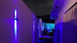 Un couloir étroit éclairé d’une lumière violette, qui mène à une pièce contenant un lit et une fenêtre.