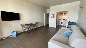 Una sala de estar moderna con un televisor de pantalla plana montado en la pared, un sofá gris con un cojín azul, una mesa de comedor con sillas, y un área de lavandería con lavadora y secadora.
