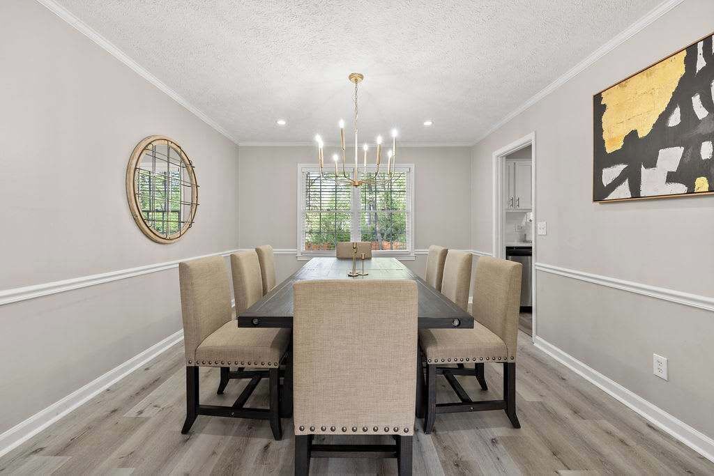 Une salle à manger avec une longue table, six chaises, un lustre, une fenêtre avec des stores et un tableau au mur.