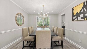 Une salle à manger avec une longue table, six chaises, un lustre, une fenêtre avec des stores et un tableau au mur.