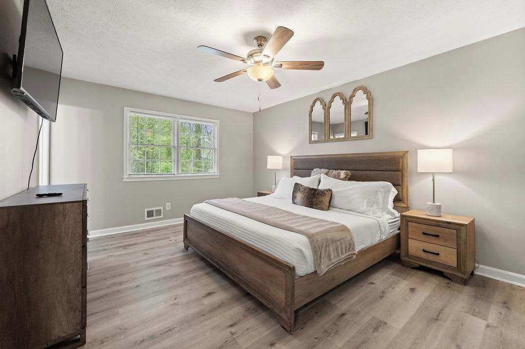 Une chambre avec un lit en bois, un ventilateur de plafond, une table de chevet avec une lampe et une fenêtre donnant sur des arbres.