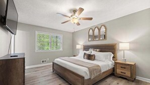 Une chambre avec un lit en bois, un ventilateur de plafond, une table de chevet avec une lampe et une fenêtre donnant sur des arbres.