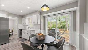Une cuisine moderne avec des meubles blancs, un coin repas avec une table ronde et des chaises, et une grande porte-fenêtre coulissante donnant sur une terrasse extérieure.