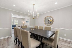 Un coin repas avec une table en bois foncé, des chaises recouvertes d’un tissu beige, un lustre et un miroir rond accroché au mur.