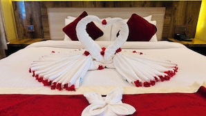 Un lit bien fait, agrémenté d’une décoration en forme de cœur réalisée avec des serviettes et des pétales de rose, avec une tête de lit en bois et un plaid rouge.