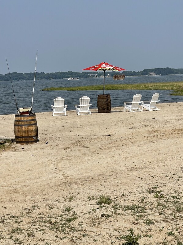 Una playa con sillas Adirondack blancas, un barril de madera y un paraguas rojo.