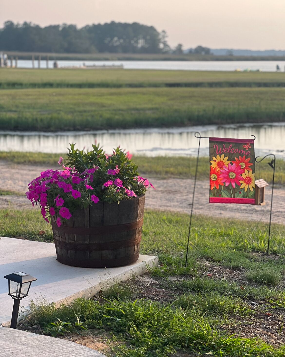 Een grote plantenbak met felroze bloemen, een bord met bloemen in verschillende kleuren en een uitzicht op een rustig wateroppervlak met een verre kustlijn.