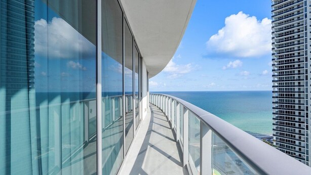 A modern balcony with glass railings overlooking a clear blue sea and a distant cityscape.