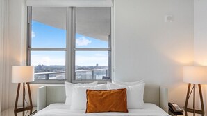 A hotel room with a large bed, two bedside lamps, a view of a cityscape through a large window, and a city bridge.