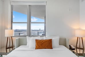 A hotel room with a large bed, two bedside lamps, a view of a cityscape through a large window, and a city bridge.