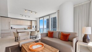 A modern apartment with a living area, kitchen, and dining space. It features a sofa, coffee table, and a view of the city through large windows.