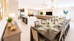 A modern kitchen with a central island, a dining area with a long table set for six, and a living area with a sofa and TV.