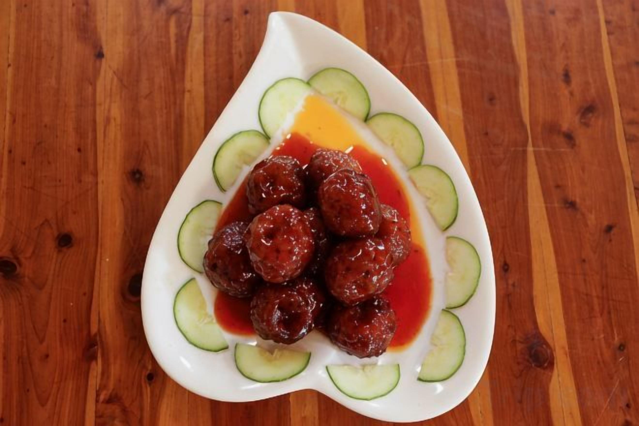 Une assiette blanche contenant des boulettes de viande et de la sauce, entourée de rondelles de concombre, posée sur une table en bois.
