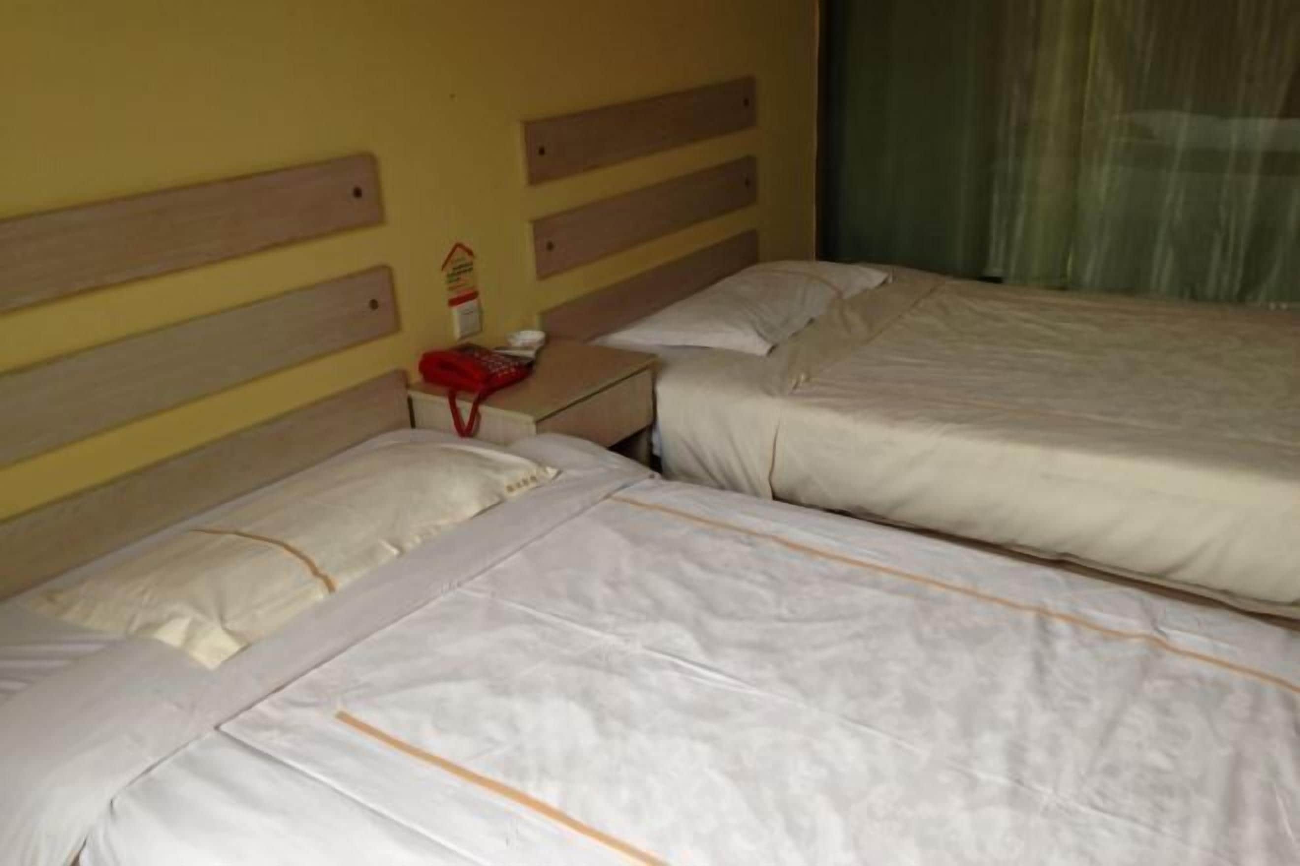 Een hotelkamer met twee eenpersoonsbedden, houten hoofdeinden, een nachtkastje met een rode telefoon en een groen gordijn.