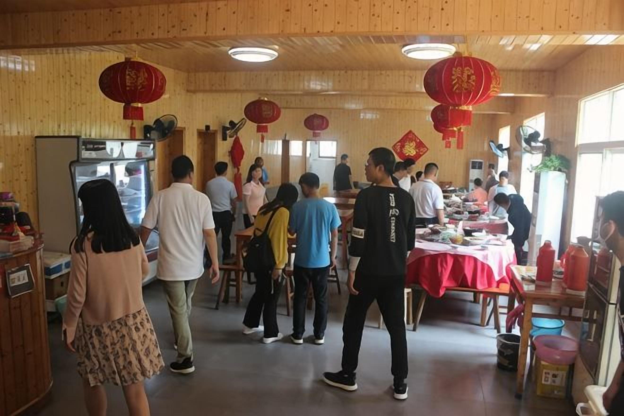 Une pièce aux murs et au plafond en bois, une longue table recouverte d’une nappe rouge, et plusieurs personnes debout autour.