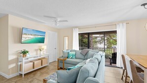 Un salon avec un canapé gris, un téléviseur fixé au mur, un ventilateur de plafond et un balcon donnant sur l’extérieur.