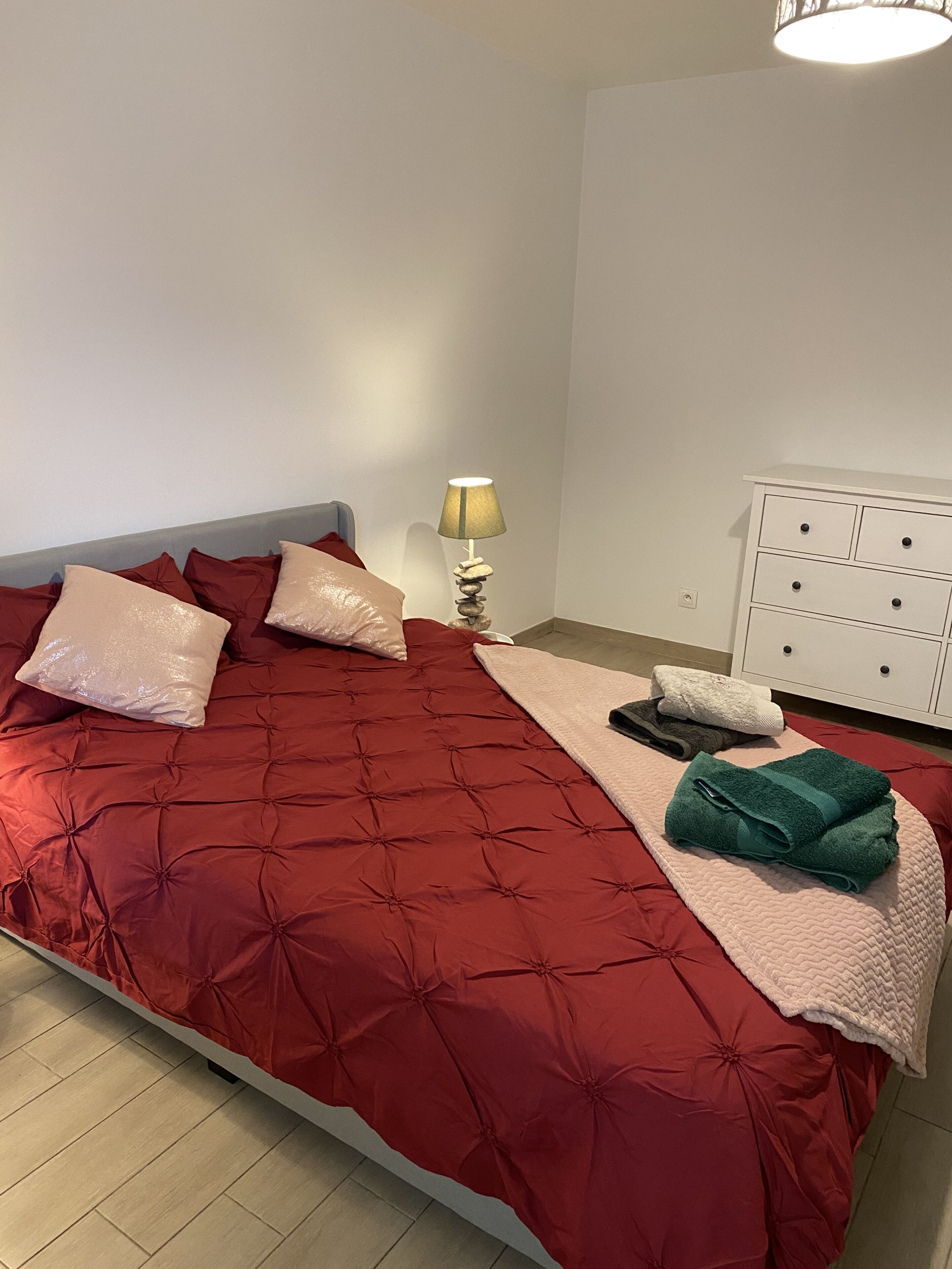 Een opgemaakt bed met een rode quilt, een bedlampje en een witte dressoir met lades.