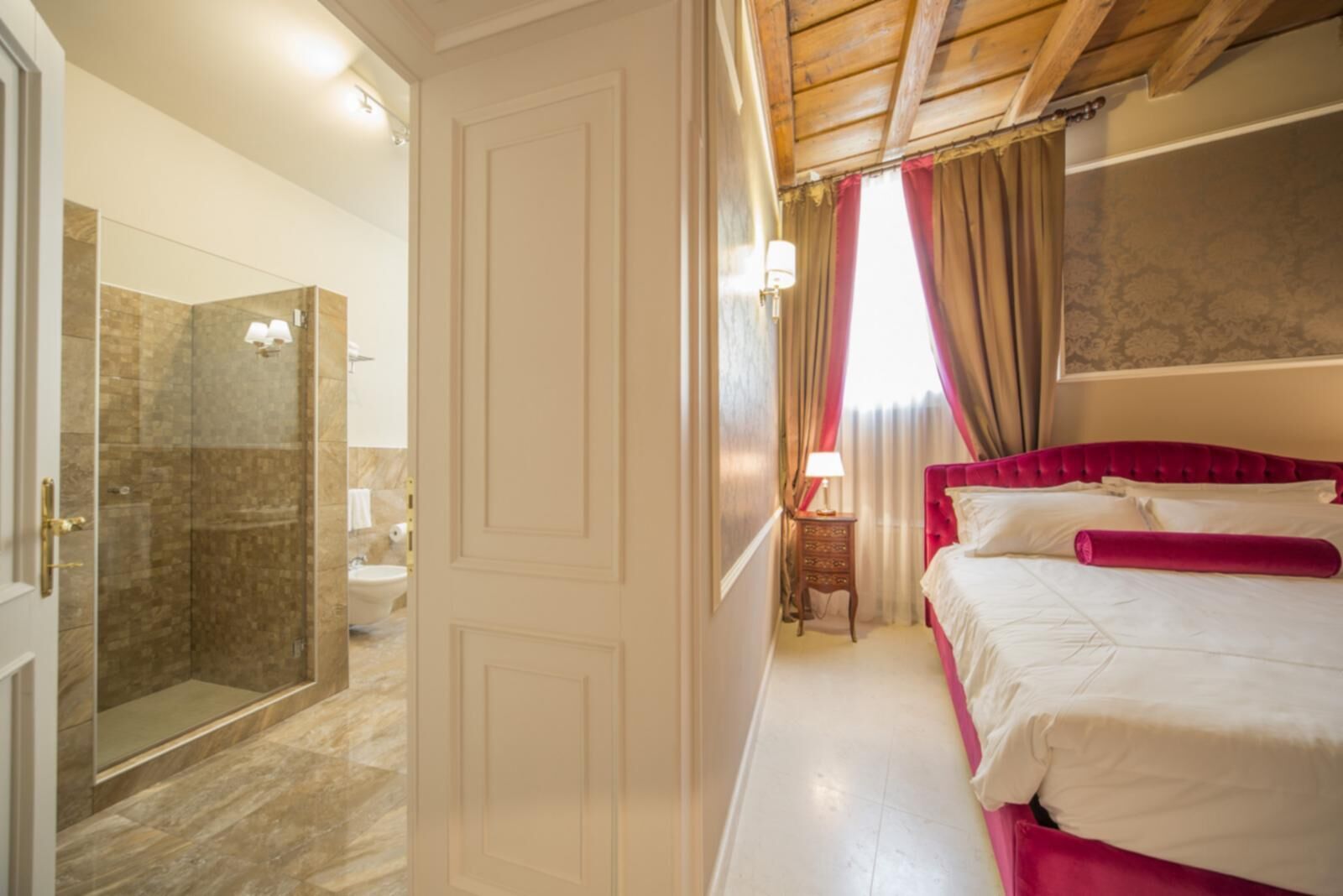 A hotel room with a glass-enclosed shower, a bed with red bedding, and a nightstand with a lamp.