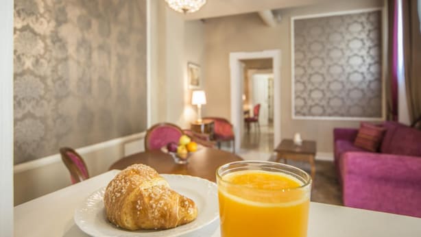 Et spisebord med en croissant og et glas appelsinjuice, en lilla sofa og en lysekrone i et hotelværelse.