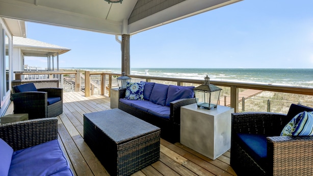 Eine überdachte Terrasse im Freien mit Korbmöbeln, einem weißen Tisch und Blick auf das Meer.