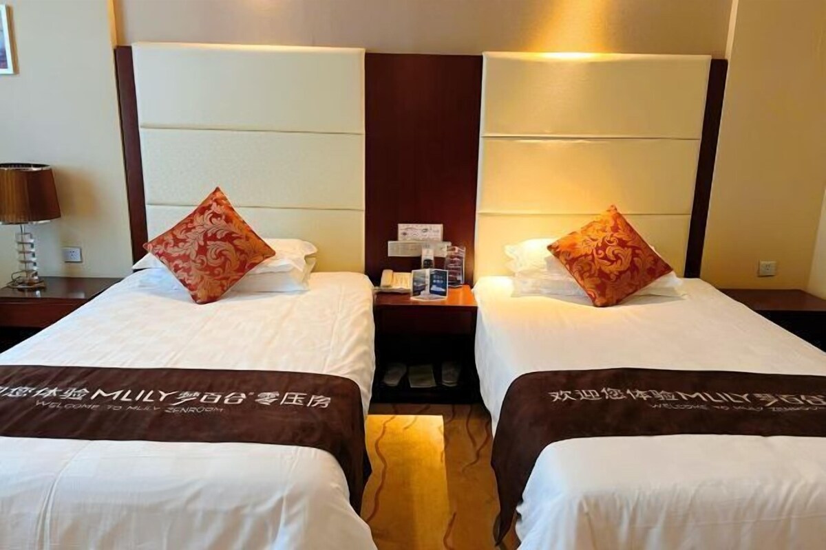 A hotel room with two beds, a nightstand, a lamp, and a wall-mounted shelf with items.