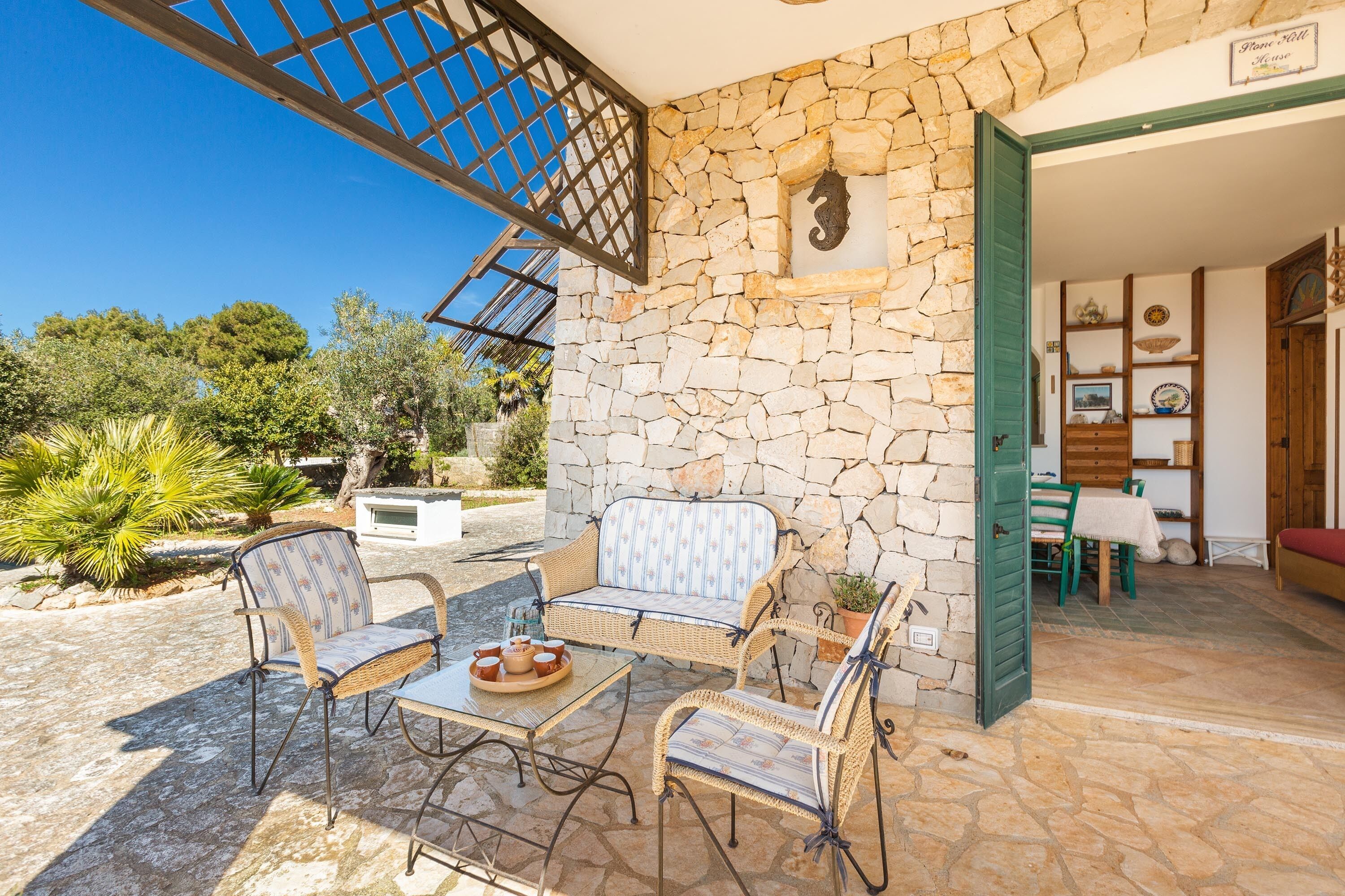 A stone-walled outdoor seating area with wicker furniture and a glass table, leading to a room with a green door and a stone fireplace.