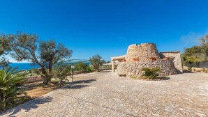A stone building with a circular tower, surrounded by a stone-paved courtyard and olive trees.