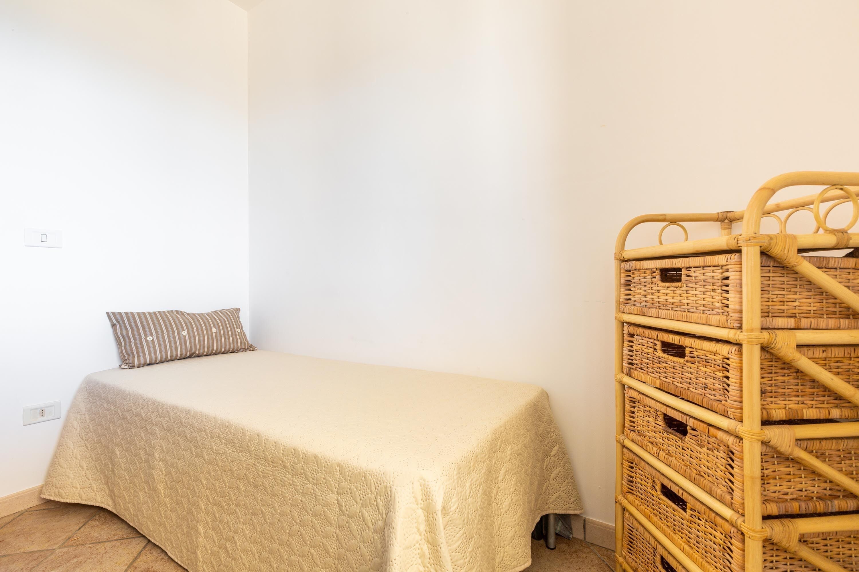 Ein kleines Zimmer mit einem Einzelbett, einer schlichten beigefarbenen Bettdecke und einem Kopfkissen. Gegen die Wand lehnt ein Korbuscheinlagerung.