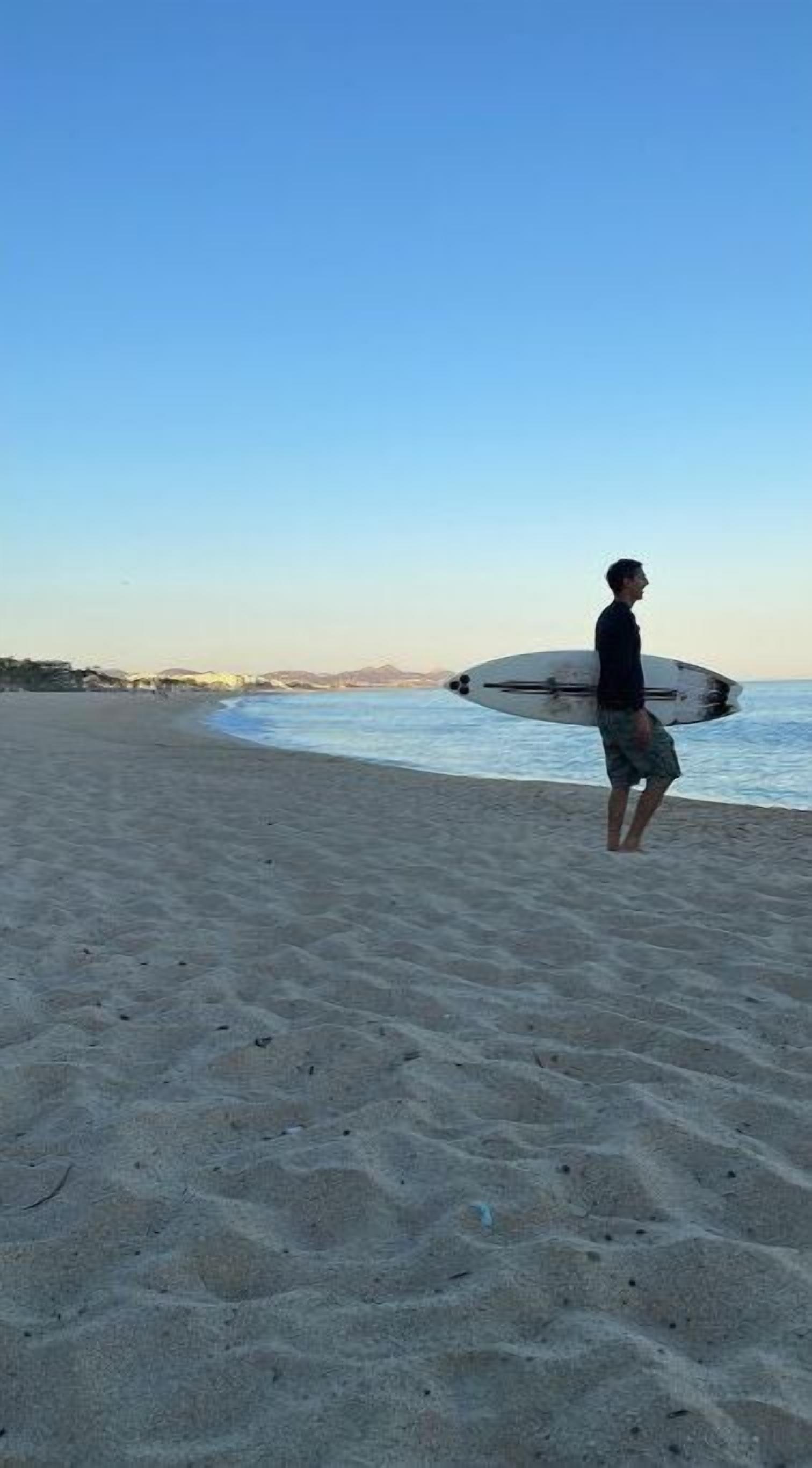 A person holding a surfboard on a sandy beach with a clear sky.
