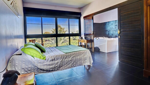 A bedroom with a large window, a bed with green pillows, a desk, and a bathroom with a bathtub. - La Villa des Mainates - Jacuzzi et vue mer (Le Tampon)