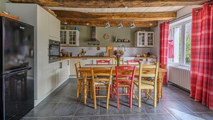 Une cuisine avec des poutres en bois, une table à manger avec des chaises, un réfrigérateur et une fenêtre avec des rideaux.