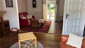 Un salon avec un canapé rouge, une table basse en bois et un tapis à motifs. On y trouve une porte avec des panneaux en verre dépoli, et de cette porte, on aperçoit un jardin.