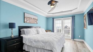 A bedroom with a bed, a nightstand, a lamp, a ceiling fan, and a window with curtains. - Whalecome to the beach-elevator (Carolina Beach)
