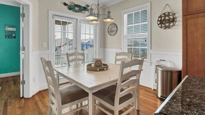 A dining area with a wooden table and chairs, a chandelier, and a view of the outside through large windows with blinds. - Whalecome to the beach-elevator (Carolina Beach)